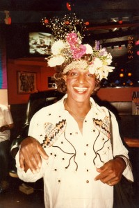 A picture of a smiling, decadently beautiful flower arrangement hat wearing Marsha "Pay It No Mind" Johnson